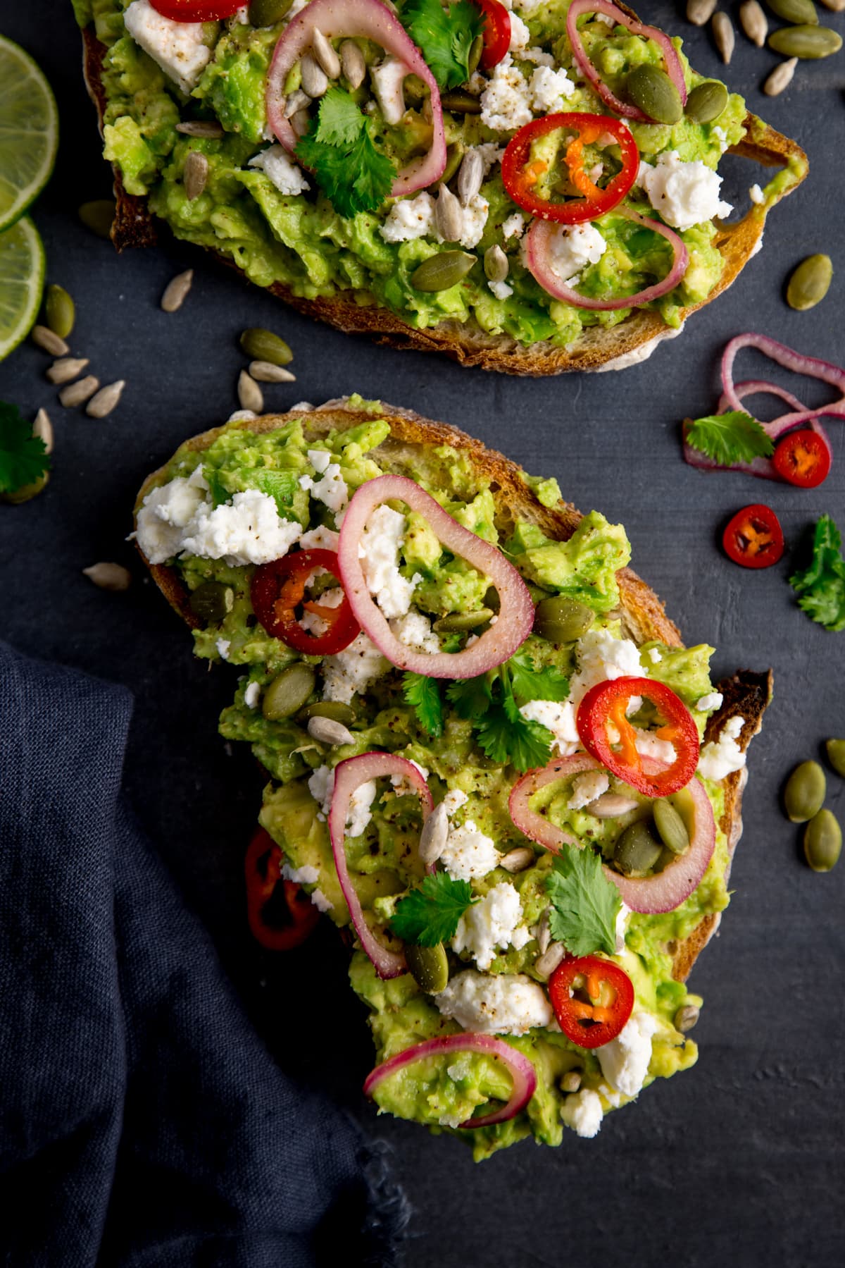 Two pieces of loaded avocado toast with feta, shallot, sliced red chilli, seeds and coriander (cilantro) on a dark grey background. There are ingredients scattered around and a dark grey napkin on the left of the frame.