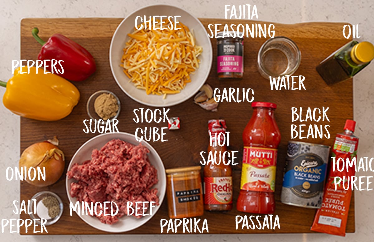 An overhead shot of the ingredients for Tex-Mex Minced Beef arranged on a wooden cutting board, which in on a white surface. Each ingredient is labelled in white and is as follows: peppers, onion, salt, pepper, sugar, minced beef, cheese, stock cube, paprika, fajita seasoning, garlic, hot sauce, passata, water, black beans, oil, and tomato puree.