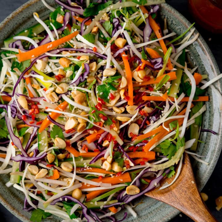 A square, closeup shot of Asian Slaw. The Asian Slaw is in a large light grey bowl, with a wooden spoon sticking out of the dish. In the top right of the image, you can slightly see a clear glass dish with the remaining dressing in it. This is all set on a dark grey background.
