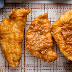 A square, overhead shot of three fish fillets, battered in the beer batter. There are three golden battered fish fillets laid side by side on a copper wire rack. They are topped with flakes of salt. The wire rack is on a white marble background, and in the top left corner, you can slightly see a blue napkin.