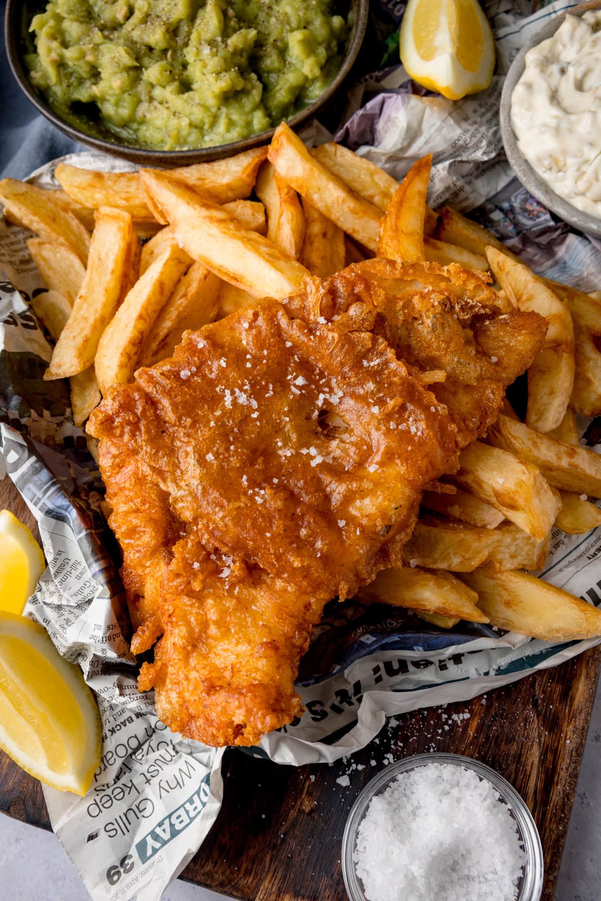 A tall, overhead shot of beer-battered fish. There is one beer-battered fish fillet, topped with flakes of salt, on a bed of chips. The food is on top of a newspaper. To the left of the fish and chips are two wedges of lemon. In the top left of the image, there is a black bowl of mushy peas, with another wedge of lemon to the right of it. In the top right of the image, there is a grey dish of tartare sauce. This is all on a wooden board. On the bottom right of the wooden board, there is a clear dish of sea salt. This is all placed on a white marble background.
