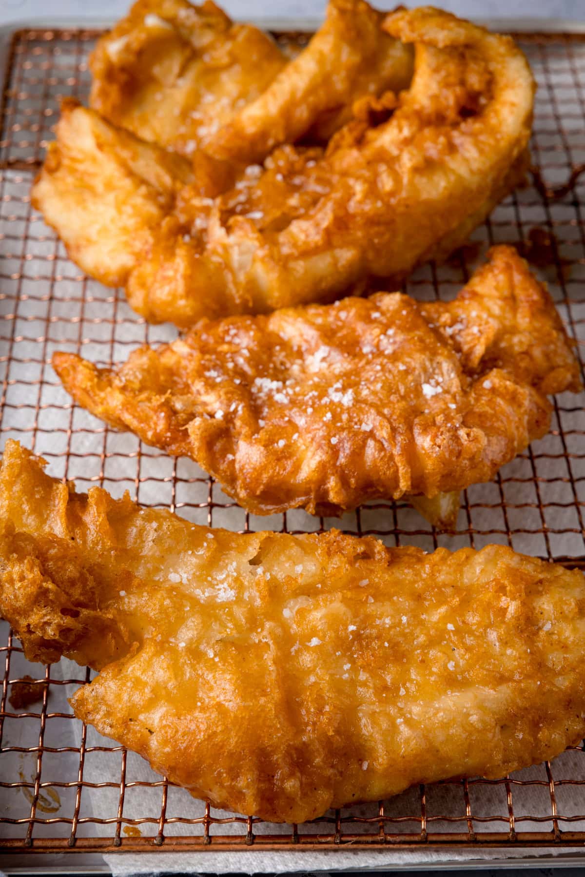 A tall shot of three fish fillets, battered in the beer batter. There are three golden battered fish fillets laid side by side on a copper wire rack. They are topped with flakes of salt. The wire rack is on a white marble background.