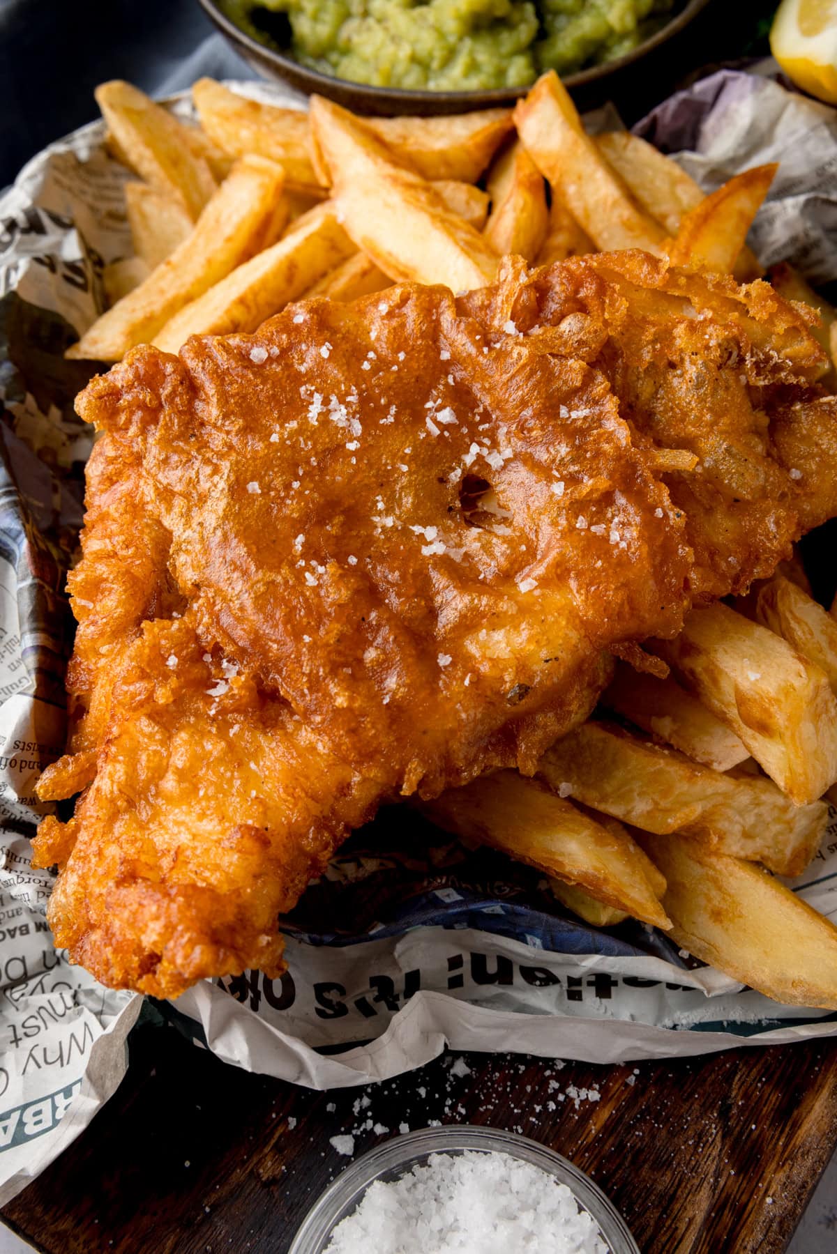 A tall, close-up shot of beer-battered fish. There is one beer-battered fish fillet, topped with flakes of salt, on a bed of chips. The food is on top of a newspaper. In the top middle of the image, there is a black bowl of mushy peas, with another wedge of lemon to the right of it. This is all on a wooden board. On the bottom middle of the wooden board, there is a clear dish of sea salt.