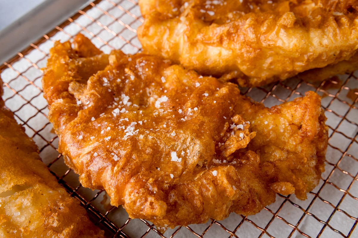 A wide, close-up shot of three fish fillets, battered in the beer batter. There are three golden battered fish fillets laid side by side on a copper wire rack. They are topped with flakes of salt. The wire rack is on a white marble background.