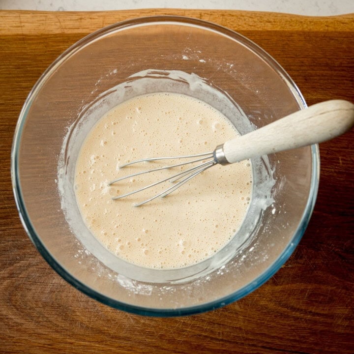 A square, overhead shot of the finished beer batter. The batter is in a clear, glass bowl with a whisk wooden handle in the batter. All this is laid on a wooden board.
