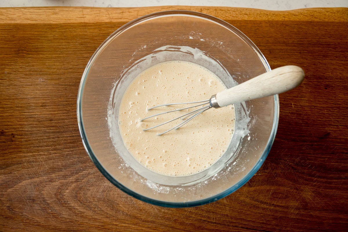 A wide, overhead shot of the finished beer batter. The batter is in a clear, glass bowl with a whisk wooden handle in the batter. All this is laid on a wooden board.