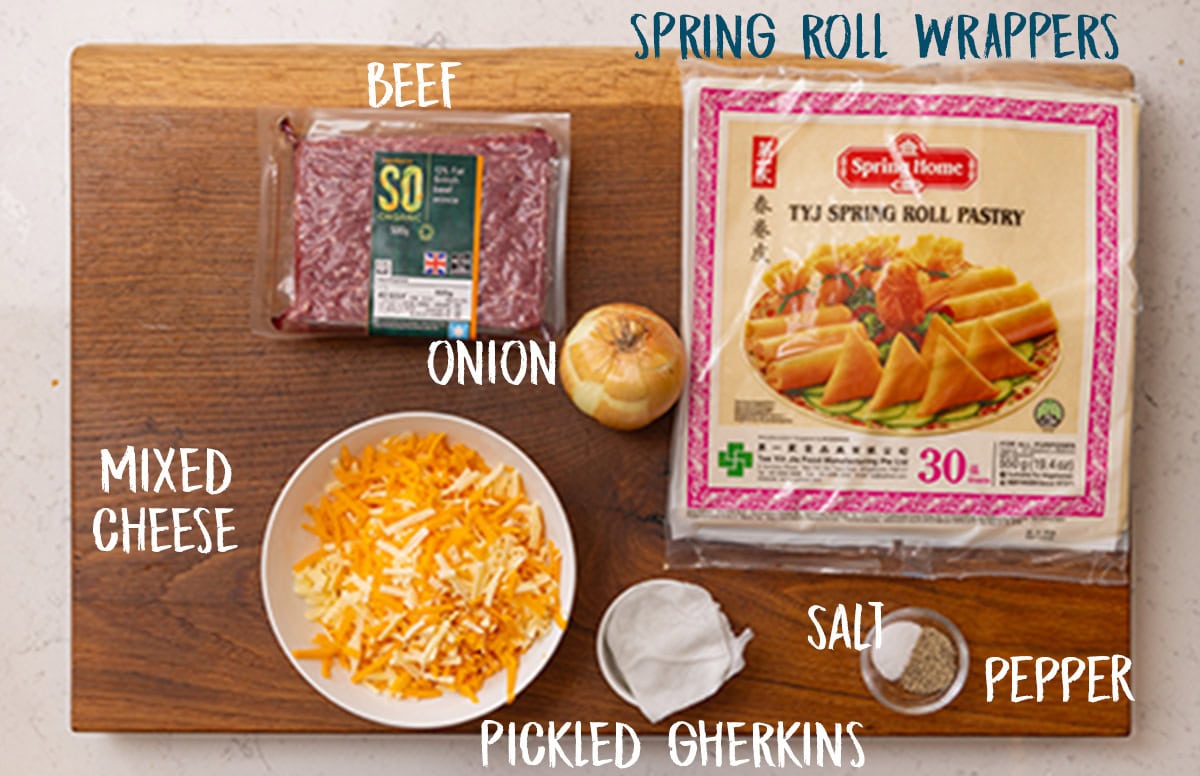 The ingredients for Cheeseburger Spring Rolls are set out on a wooden cutting board. They are each labelled in white text, except the spring roll wrappers are labelled in navy blue. The ingredients are as follows: Beef, Onion, Mixed Cheese, Pickled Gherkins, Salt, Pepper, and Spring Roll Wrappers.