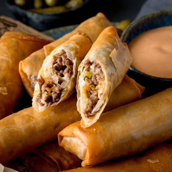 A square shot of Cheeseburger Spring Rolls. There are 6 whole spring rolls stacked horizontally next to each other, and on top of the stack (in the middle of the image) there is another spring roll that has been cut in half and the two halves are arranged next to each other so you can see the filling. This is set on a wooden board. In the right of the background, you can see a dark blue dish filled with burger sauce. And in the top left of the background, you can slightly see another blue dish with pickled gherkins in it. This is all set on a navy blue background.