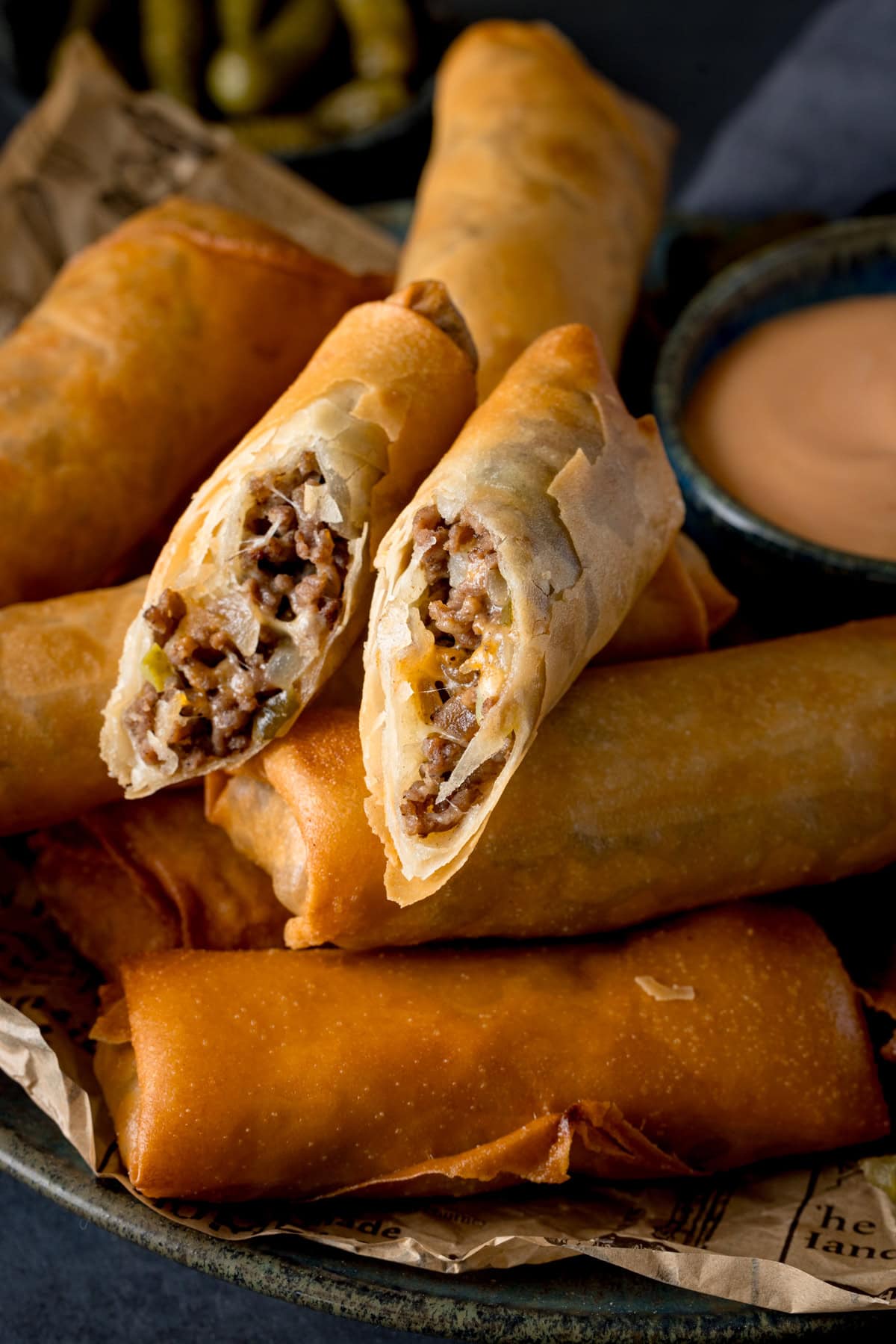 A tall shot of Cheeseburger Spring Rolls. There are 6 whole spring rolls stacked horizontally next to each other, and on top of the stack (in the middle of the image) there is another spring roll that has been cut in half and the two halves are arranged next to each other so you can see the filling. This is set on a wooden board. In the right of the background, you can see a dark blue dish filled with burger sauce. And in the top left of the background, you can slightly see another blue dish with pickled gherkins in it. This is all set on a navy blue background.