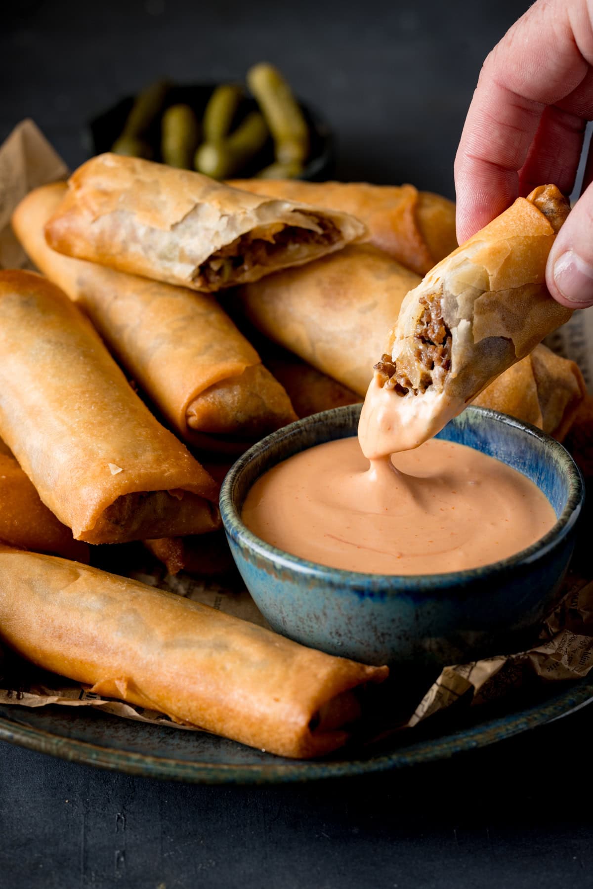 A tall shot of Cheeseburger Spring Rolls. There are 6 whole spring rolls stacked horizontally next to each other, and on top of the stack (in the middle of the image) there is another spring roll that has been cut in half and one of the halves is arranged so you can see the filling. The other half is being held by a hand on the right of the screen and is being dipped into a dark blue dish, filled with burger sauce. The dark blue dish with burger sauce is on the same dish as the spring rolls, and the spring rolls are stacked around it. This is all set on a dark blue plate, which is lined with a brown newspaper. And in the top left of the background, you can slightly see another blue dish with pickled gherkins in it. This is all set on a navy blue background.