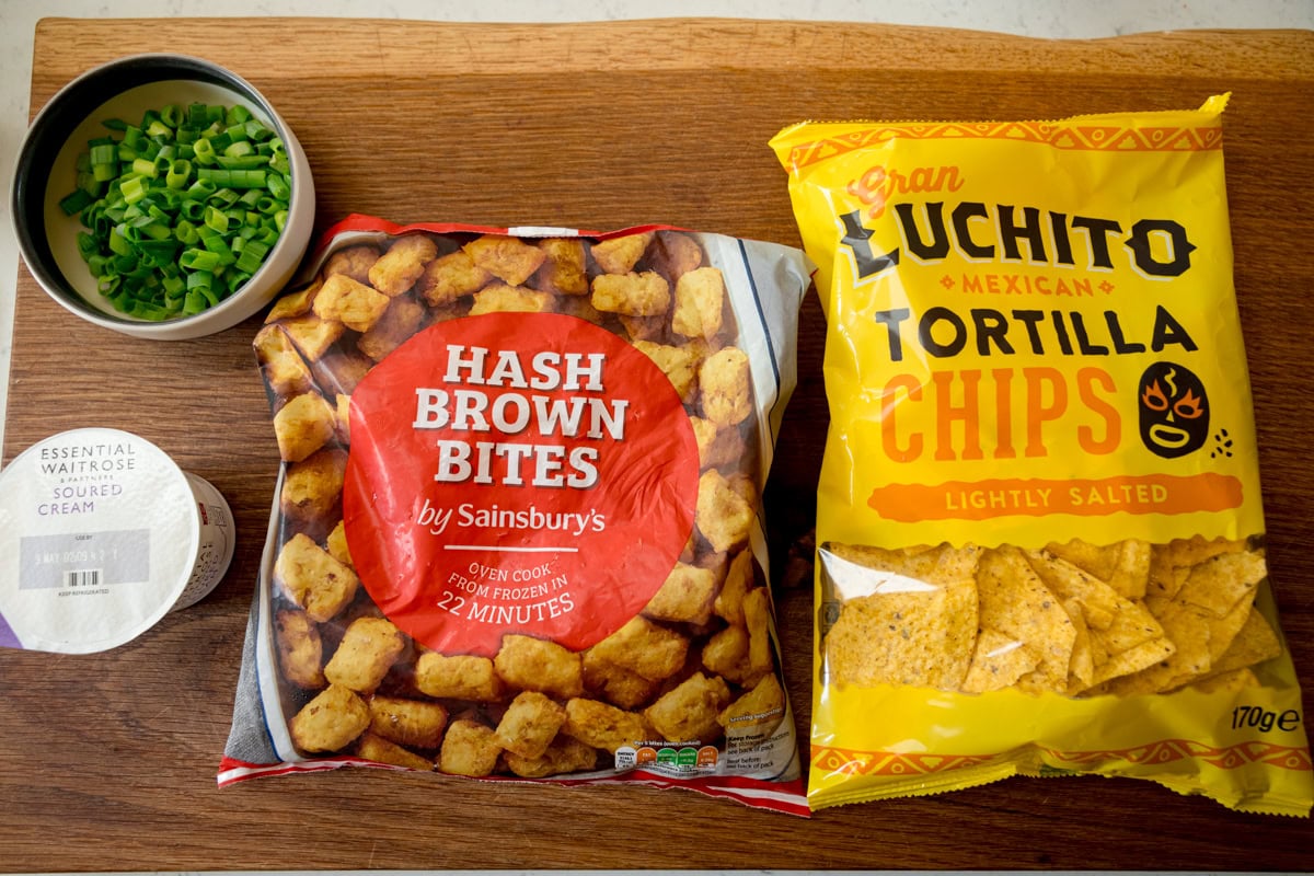 The ingredients for the Loaded Tocho fixings are laid out on a wooden board. They are as follows: Tortilla Chips, Hash Brown Bites, Soured Cream and Spring Onions.