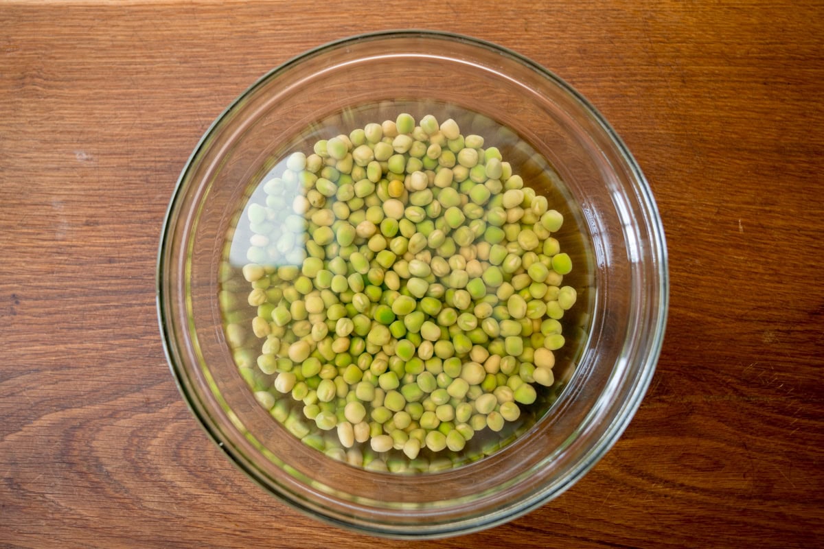 A wide overhead image of some dried peas soaking in water, in a clear glass bowl. This is on a wooden background.