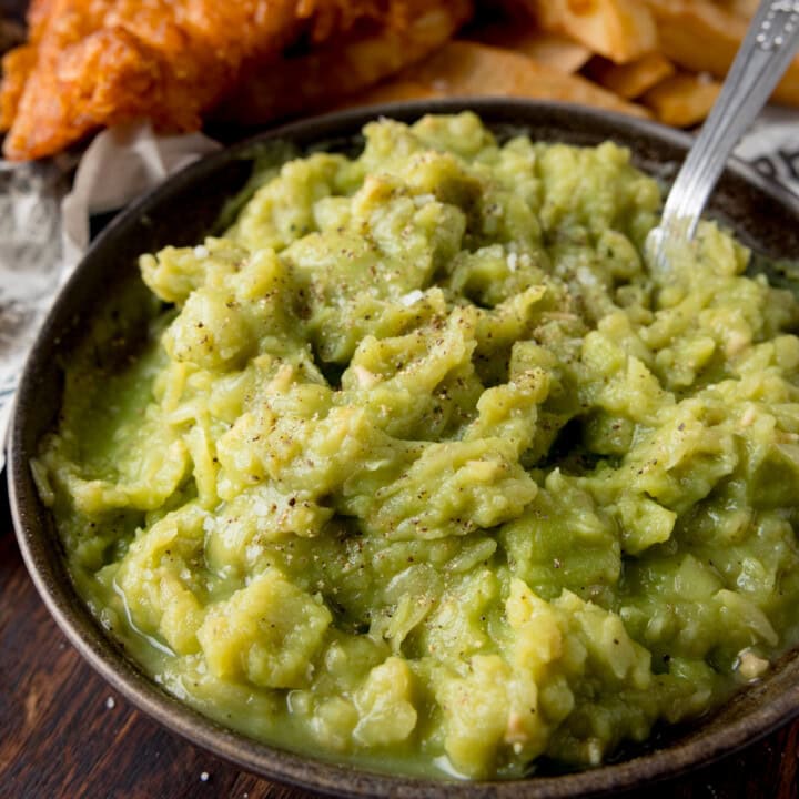 A square image of mushy peas in a black bowl, with a silver spoon sticking out of it. In the background you can see chip shop style fish and chips on some newspaper. This is on a dark wooden board.