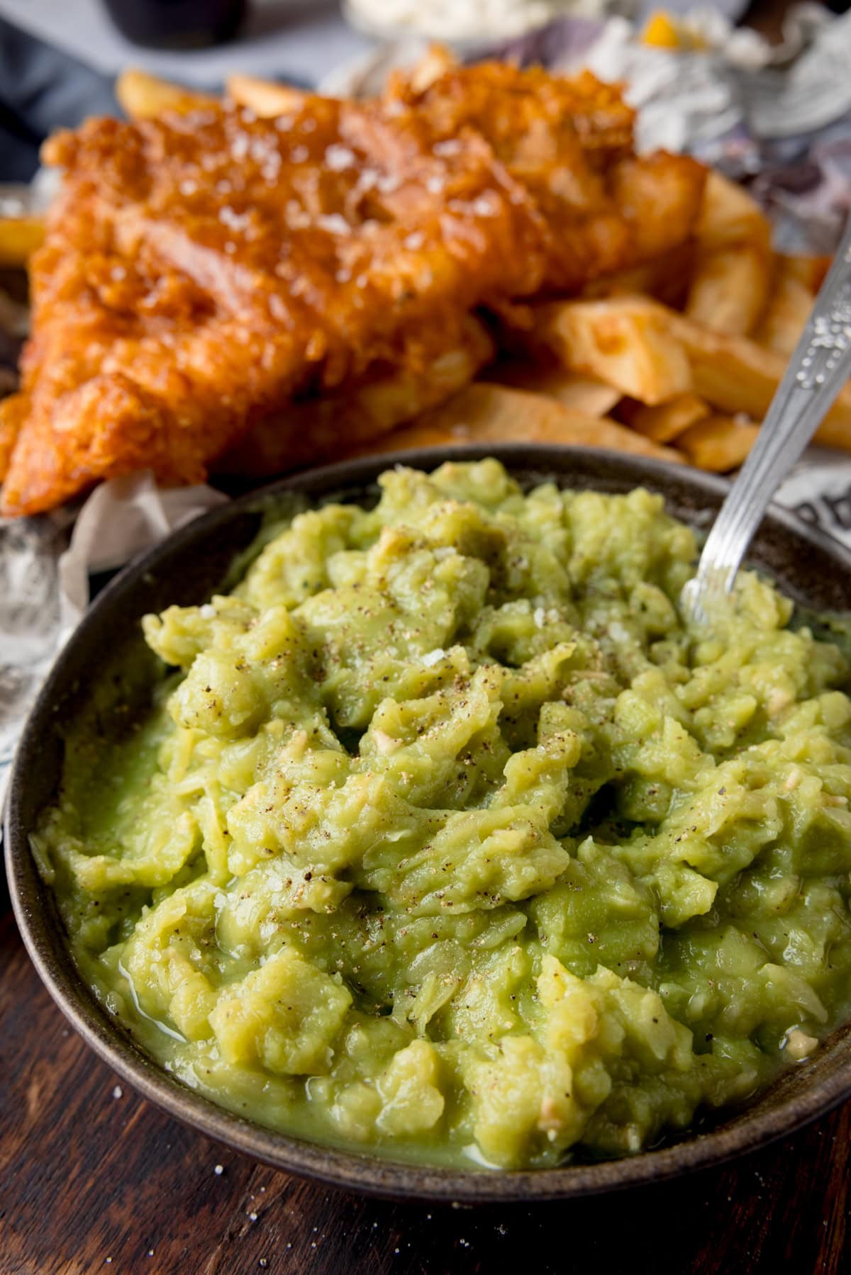 A tall image of mushy peas in a black bowl, with a silver spoon sticking out of it. In the background you can see chip shop style fish and chips on some newspaper. This is on a dark wooden board.