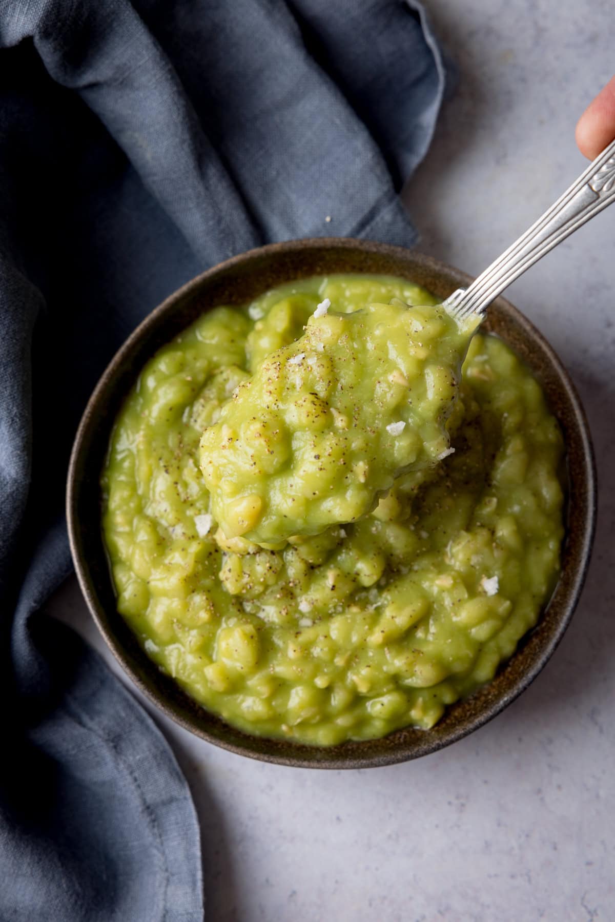 A tall, overhead shot of Chip Shop Style Mushy Peas in a black bowl. There is a silver spoon, sticking out of the right of the dish. A slate grey napkin covers the left of the background. The background is light grey.