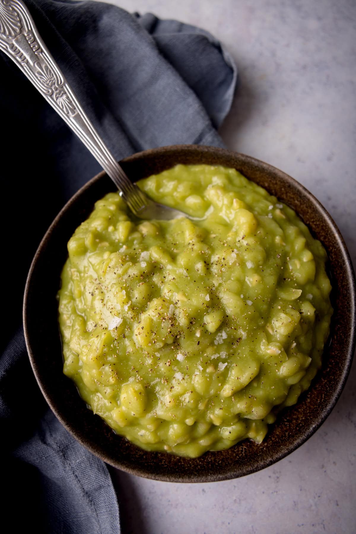 A tall, overhead shot of Chip Shop Style Mushy Peas in a black bowl. There is a silver spoon, sticking out of the left of the dish. A slate grey napkin covers the left of the background. The background is light grey.