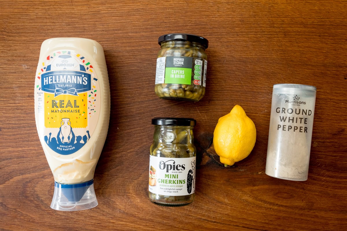 A wide overhead shot of the ingredients for tartare sauce are laid out on a wooden board. They are as follows: mayonnaise, mini gherkins, capers in brine, a lemon, and ground white pepper.