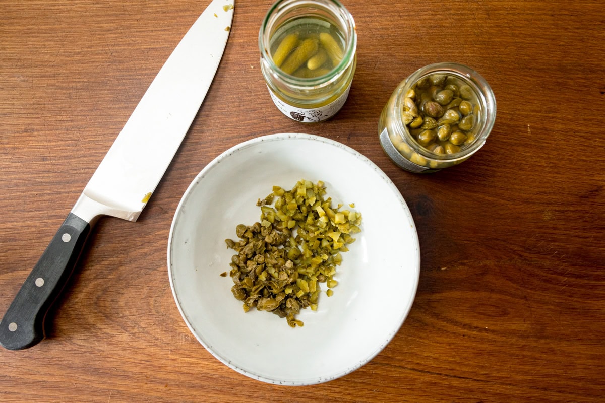 A wide overhead shot of the prep for tartare sauce. There is a white bowl which has some finely chopped pickled gherkins and capers. To the left of the bowl, there is a large silver knife with a black handle. To the top middle and left of the bowl, there is the open jars of pickled gherkins and capers. This is all on a wooden board.