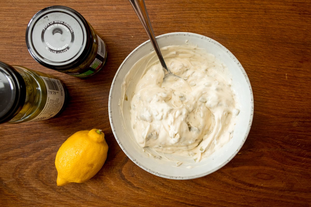 A wide overhead shot of the prep for tartare sauce. In the middle of the image, there is a white bowl with the completed tartare sauce in it, with a silver spoon sticking out of it. To the right of the bowl, there are closed jars of pickled gherkins and capers and a whole lemon. This is on a wooden board.