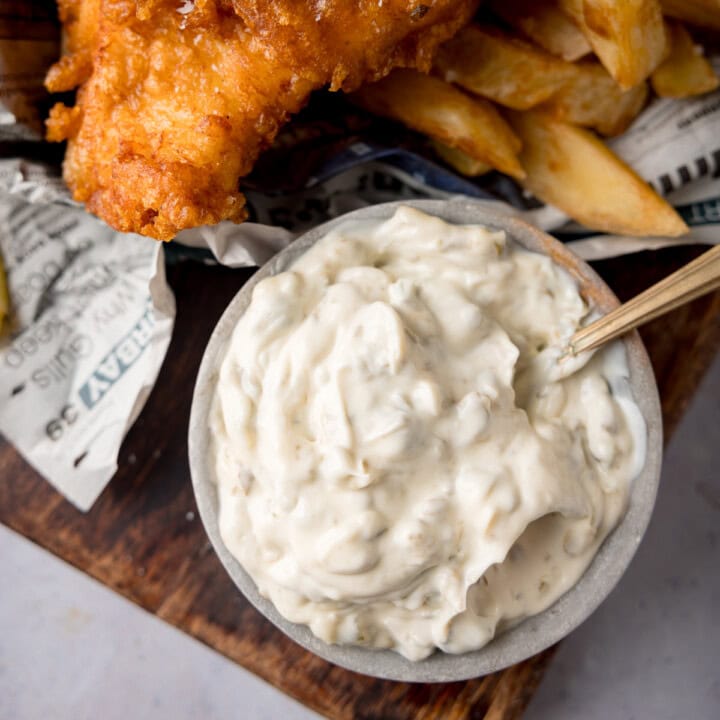 A square, overhead shot of Chip Shop-Style Tartare Sauce. In the centre of the image, there is a light grey bowl of tartare sauce with a gold spoon sticking out of the left of the bowl. In the top of the background, there are some Chip Shop-Style Fish and Chips on some newspaper. The food is placed on a dark wooden board. This is all on a light grey background.