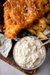 A tall, overhead shot of Chip Shop-Style Tartare Sauce. In the centre of the image, there is a light grey bowl of tartare sauce with a gold spoon sticking out of the left of the bowl. In the top of the background, there are some Chip Shop-Style Fish and Chips on some newspaper. The food is placed on a dark wooden board. This is all on a light grey background.