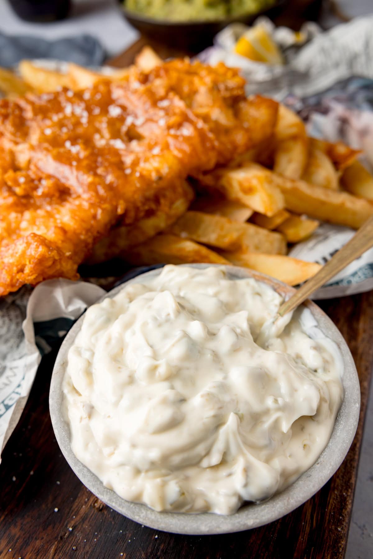 A tall shot of Chip Shop-Style Tartare Sauce. In the centre of the image, there is a light grey bowl of tartare sauce with a gold spoon sticking out of the left of the bowl. In the top of the background, there are some Chip Shop-Style Fish and Chips on some newspaper. The food is placed on a dark wooden board.