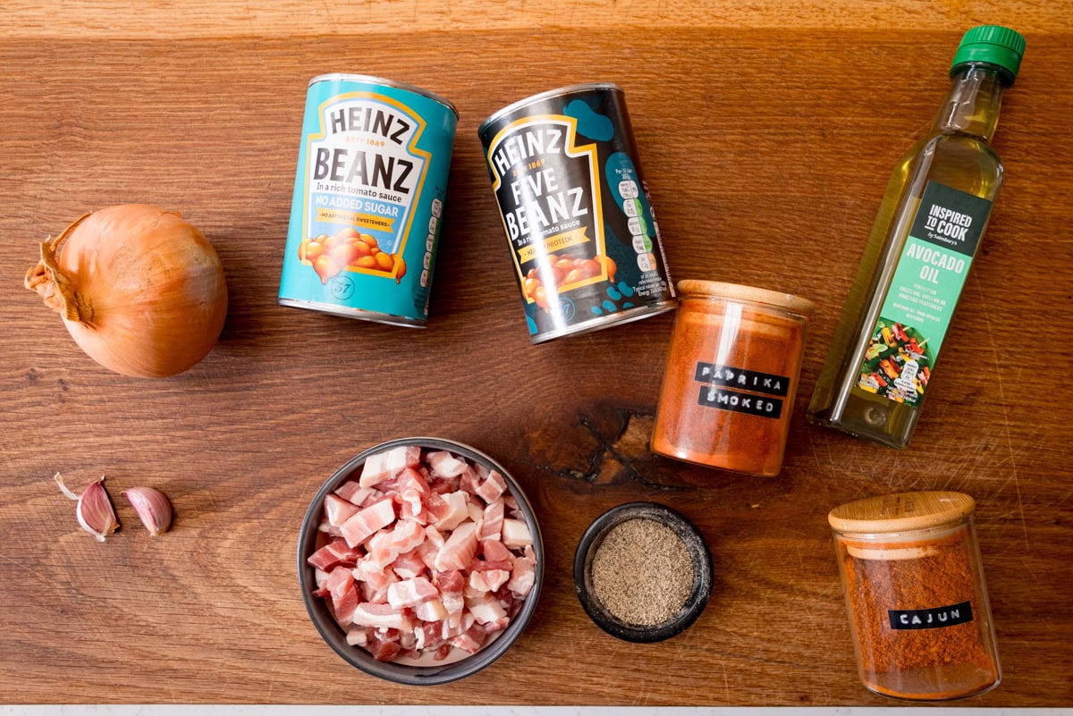 An overhead shot of the ingredients for Easy BBQ Beans. The ingredients are laid out on a wooden cutting board. They are as follows: an onion, 2 cloves of garlic, beans in tomato sauce, 5 means in tomato sauce, bacon lardons, smoked paprika, ground black pepper, avocado oil and cajun seasoning.