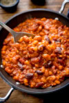 A tall shot of BBQ Beans. The BBQ Beans are in a black skillet, with a silver spoon sticking out of the dish. In the top of the background, you can see two small black dishes, one with salt and one with ground pepper. This is all set on a wooden background.