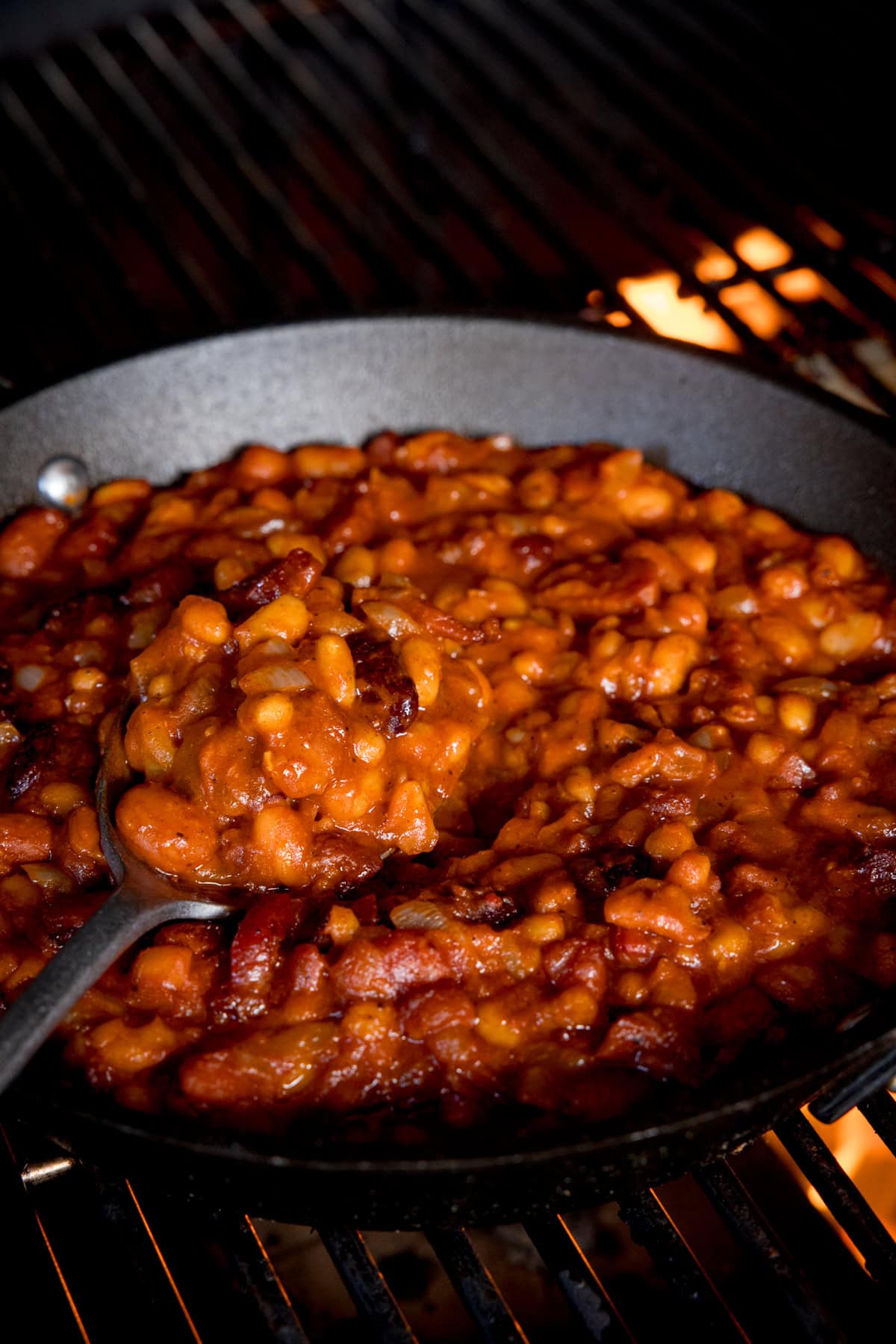 A square shot of BBQ Beans. The BBQ Beans are in a black skillet, with a silver spoon sticking out of the dish, which is scooping some of the beans in the dish. In the background, you can see that the skillet is on a lit barbeque, in the top right of the background you can see a small flame from the barbeque.