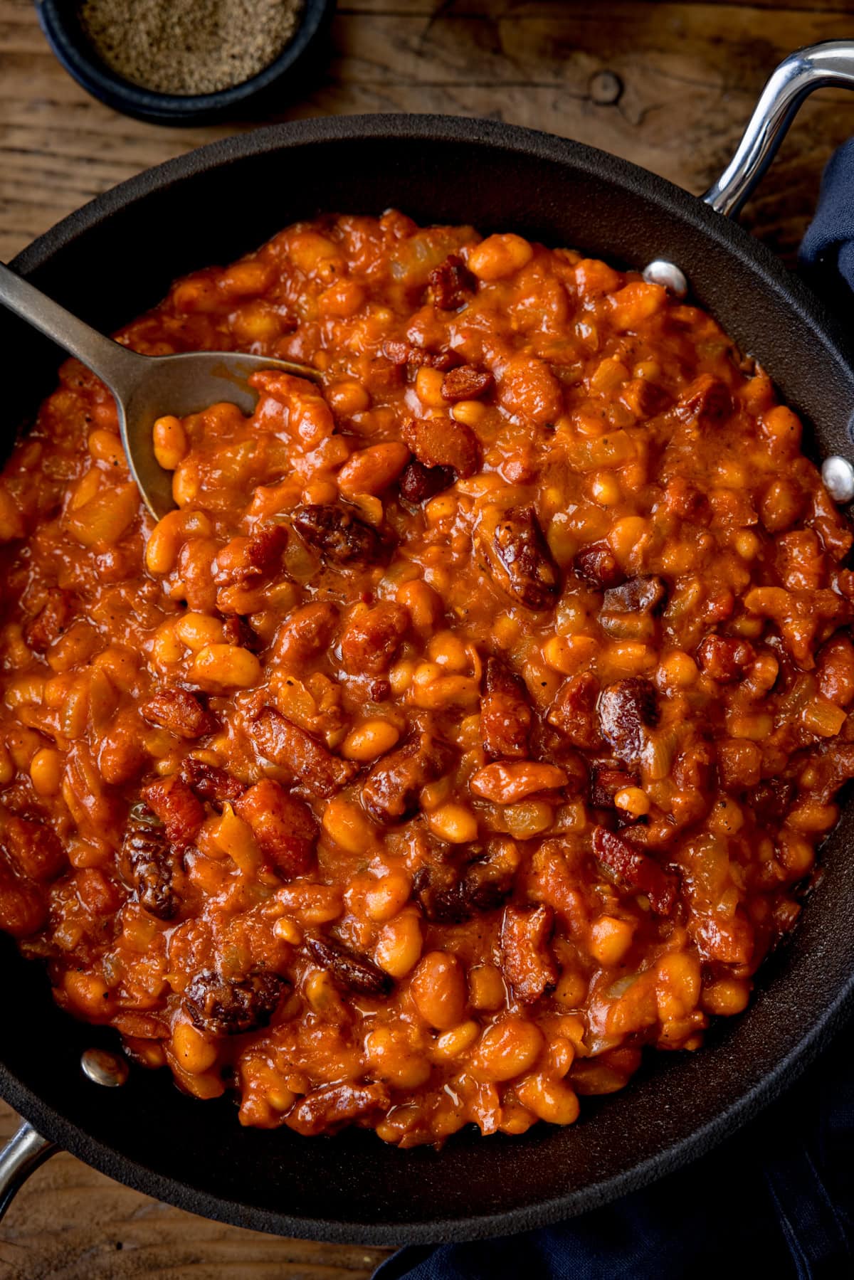 A tall, overhead shot of BBQ Beans. The BBQ Beans are in a black skillet, with a silver spoon sticking out of the dish. In the top of the background, you can see two small black dishes, one with salt and one with ground pepper. This is all set on a wooden background.