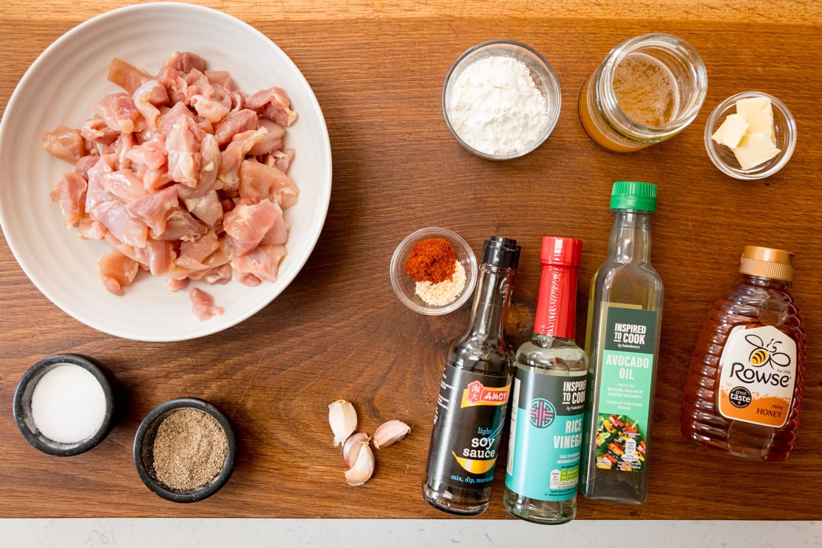 A wide, overhead shot of the ingredients for Baked Honey Garlic Chicken. They are laid out on a wooden cutting board. They are as follows: chicken, salt, pepper, cornflour, garlic powder, paprika, garlic, chicken stock, light soy sauce, rice vinegar, avocado oil, butter, and honey.