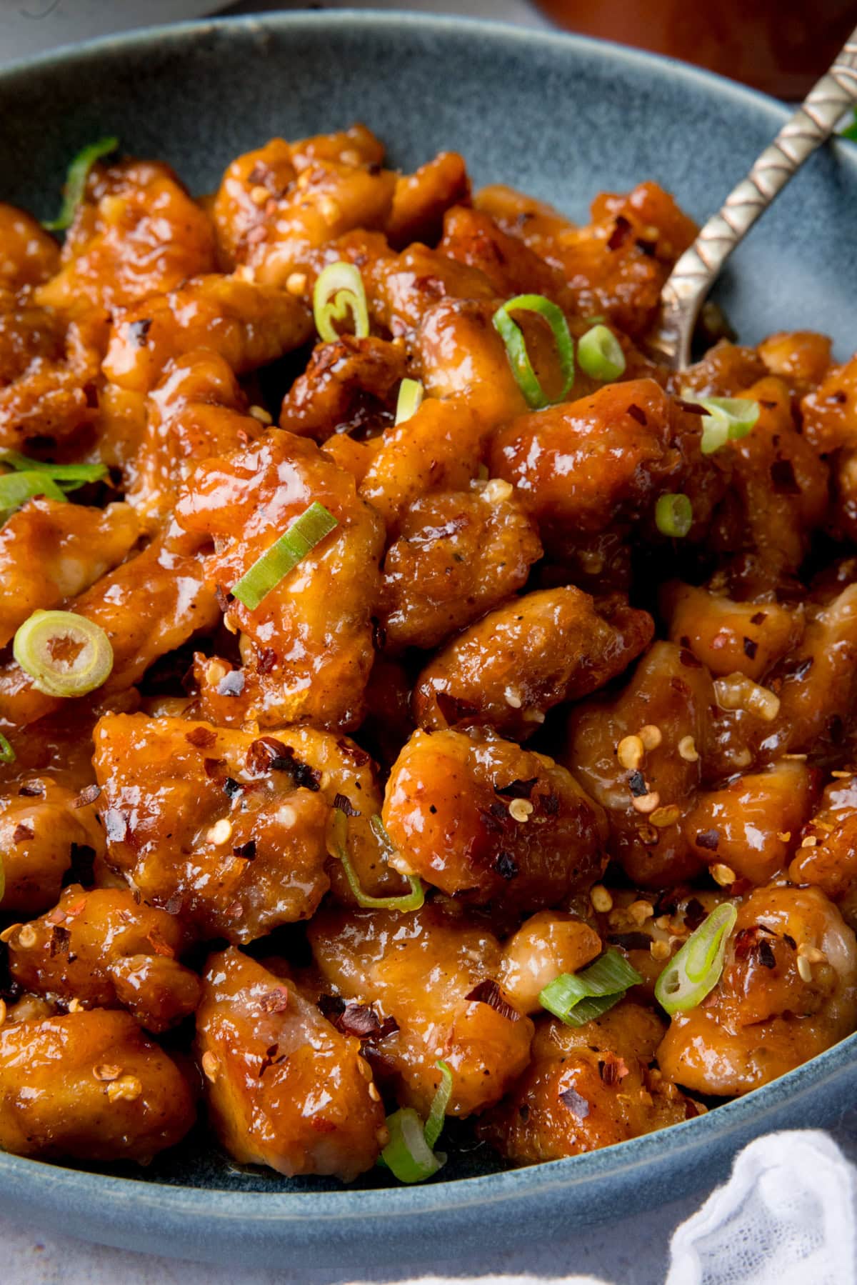 A tall, closeup shot of Baked Honey Garlic Chicken. The picture is of a blue bowl filled with chicken, which fills the image. The chicken is topped with chopped spring onion and dried chilli flakes. There is a silver spoon sticking out of the right of the bowl. In the bottom right corner, you can slightly see the edge of a white napkin.