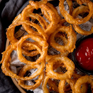 A square, overhead image of Fried Onion Rings. The onion rings are set on a brown plate, lined with black and white newspaper. To the right of the image, on the plate with the onion rings, there is a black dish filled with tomato ketchup. You can see to the left of the image, that a slate grey napkin is tucked around the plate, so the background is not visible.