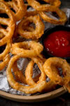 A tall, closeup image of Fried Onion Rings. The onion rings are set on a brown plate, lined with black and white newspaper. To the right of the image, on the plate with the onion rings, there is a black dish filled with tomato ketchup. This is set on a dark grey surface.