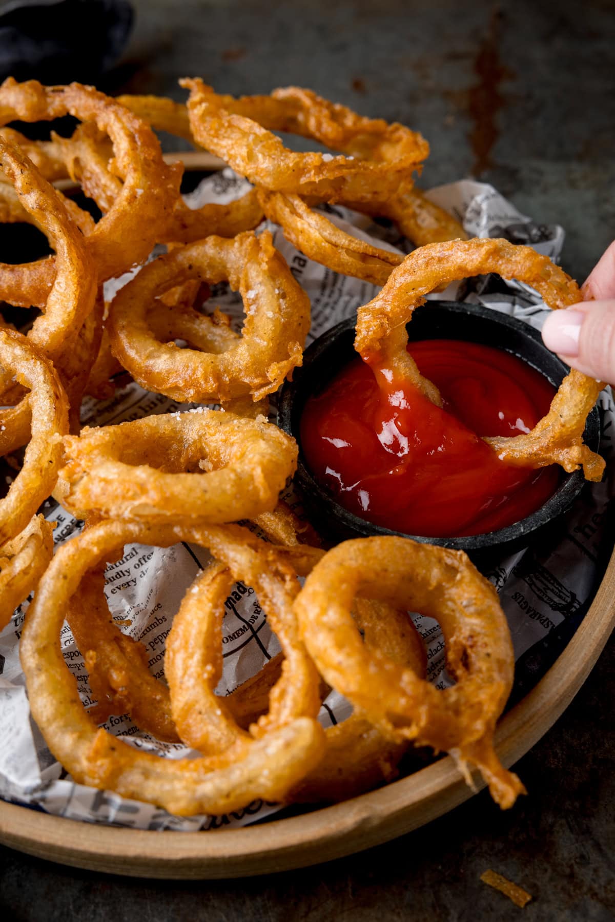 A tall image of Fried Onion Rings. The onion rings are set on a brown plate, lined with black and white newspaper. To the right of the image, on the right side of the plate with the onion rings, there is a black dish filled with tomato ketchup. Coming from the right side of the image there are two fingers holding an onion ring, which is being dipped in the ketchup pot. This is all set on a dark grey, metal background.