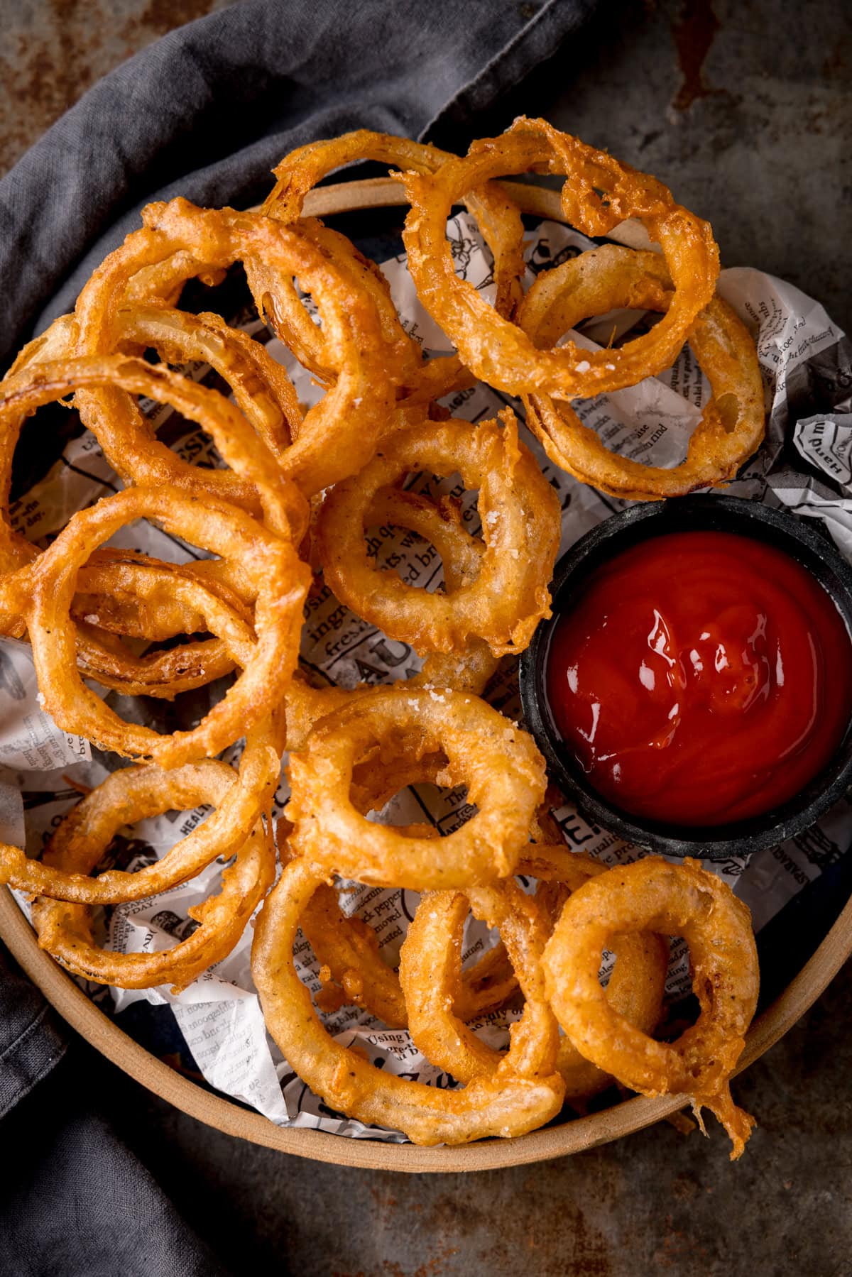 A tall, overhead image of Fried Onion Rings. The onion rings are set on a brown plate, lined with black and white newspaper. To the right of the image, on the right side of the plate with the onion rings, there is a black dish filled with tomato ketchup. You can see to the left of the main plate, that a slate grey napkin is tucked around the plate. This is all set on a dark grey, metal background.